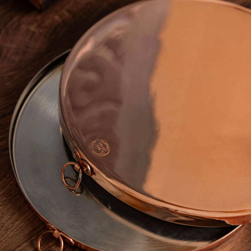 A round copper baking tray with a tin lining, displayed on a wooden surface, with a decorative hook attached to the side.